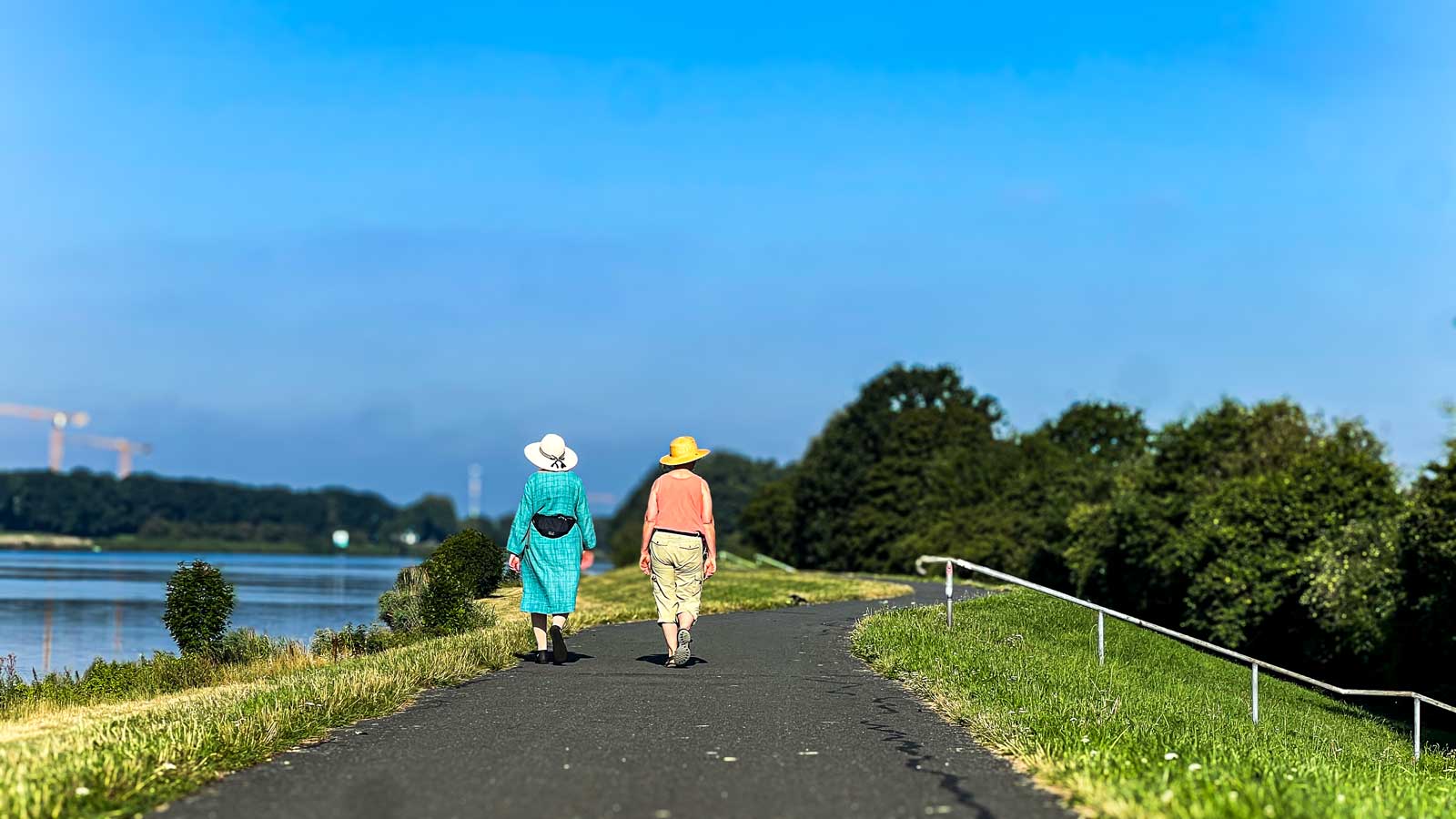 Zwei Personen mit Sonnenhut gehen entlang der Kleinen Weser auf dem Deich entlang.